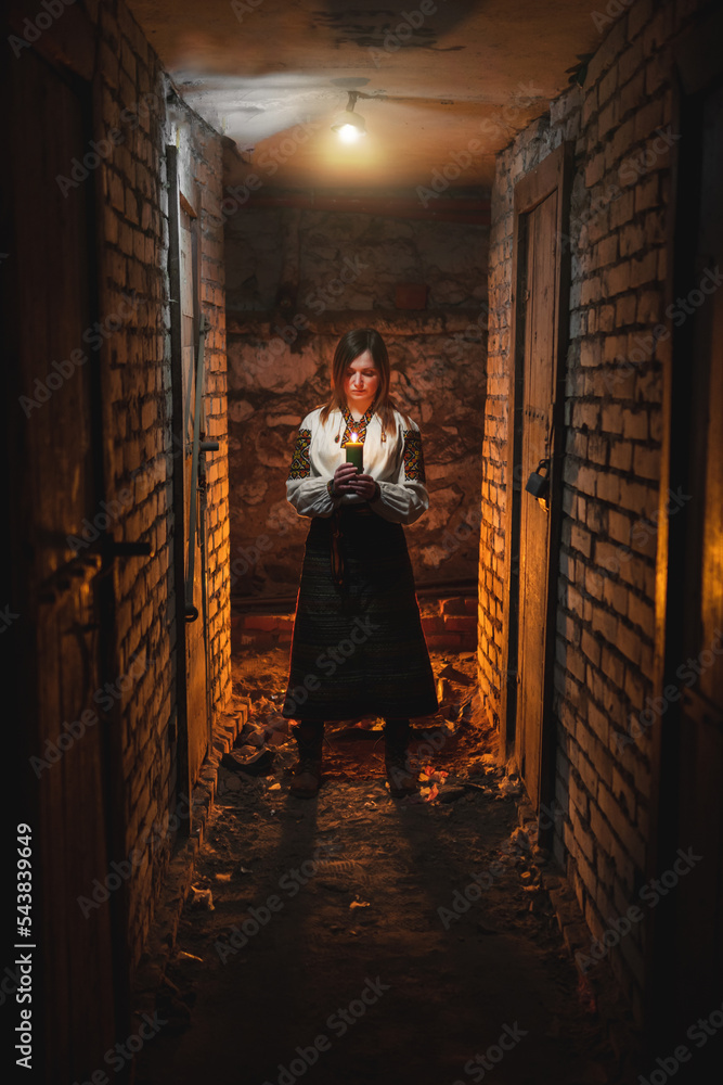 A woman in traditional Ukrainian clothing stands in a dark basement with a candle in her hands. The concept of a tragedy in Ukraine, war of shelling. Vertical photo, film noise.