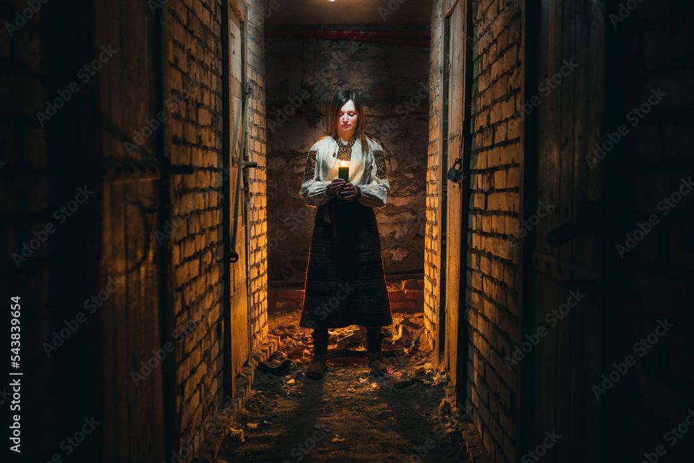 A girl, a woman in traditional Ukrainian clothing stands in a dark basement with a candle in her hands. The concept of a tragedy in Ukraine, war of shelling, blackout of electricity, light. noise 