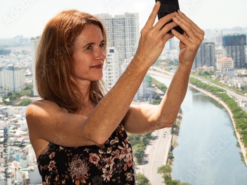 Ccaucasian woman is taking a selfie at a window with aerial view of Ho Chi Minh City, Vietnam photo