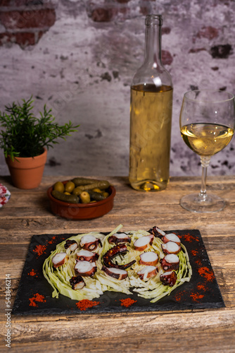 Dried octopus leg cooked on the grill and cut into pieces on a bed of shredded cabbage leaves on a stone plate  next to some olives and a glass of white wine on an old wooden table.