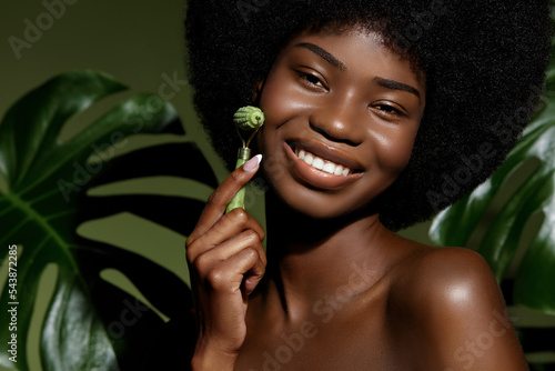 Face care Gua Sha roller. African American young woman massaging her face skin with jade roller against green leaf background. Anti age and skin tune devices.