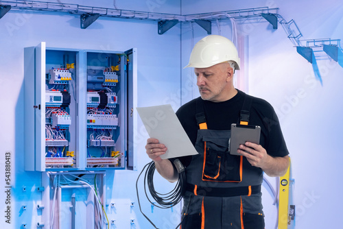 Man next to electrical panel. Electrician at work. Guy with tablet fixes electrical equipment. Man electrician reads documentation from electrical equipment. Power shield with wires fixed on wall photo