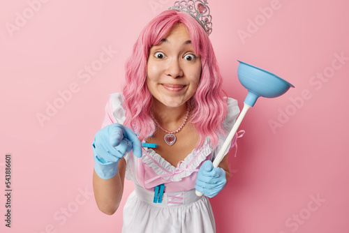 Happy surprised Asian housemaid with long pink hair holds plunger cleans everything at home wears white dress rubber blue gloves points directly at camera notices something amazing in front.