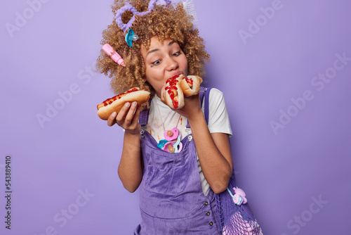 Photo of hungry pregnant woman bites delicious appetizing hot dog has unhealthy nutrition bad eating habits dressed in overalls has curly hair with different items in it isolated on purple wall