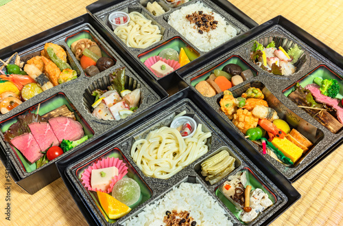 Deluxe Japanese Makunouchi bento boxes with rice,udon noodles, assortment of tsukemono preserved and nimono simmered vegetables, roast beef slices, sashimi, tofu, wagashi confectioneries on a tatami.