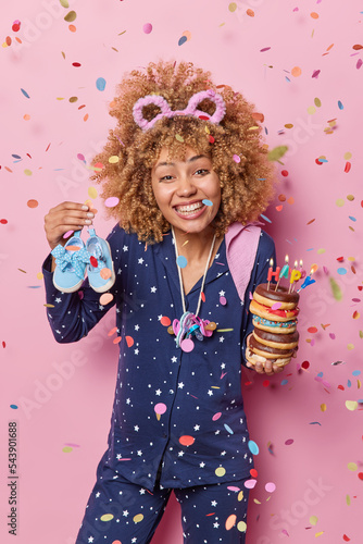 Happy pregnancy concept. Positive curly haired woman celebrates birthday at home holds shoes and pile of chocolate doughnuts dressed in comfortable pajama isolated on pink background confetti around