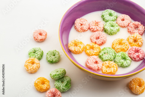 Uma vasilha com leite e cereais coloridos, e alguns cereais caídos sobre uma mesa. photo