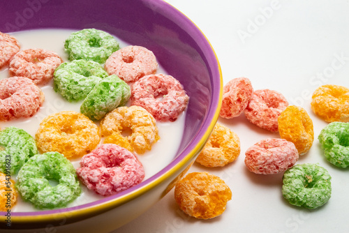 Uma vasilha com leite e cereais coloridos, e alguns cereais caídos sobre uma mesa. photo