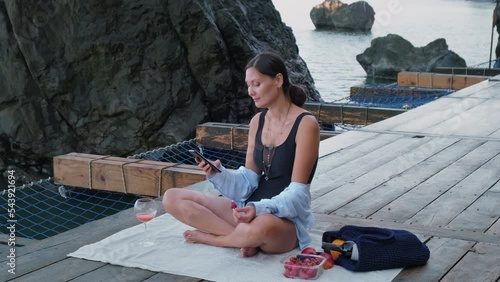 Woman Using Smartphone and Relaxing at the Sea. Female Wrights Messages on the Phone and Enjoying with Glass of Juice on the Rocky Beach.