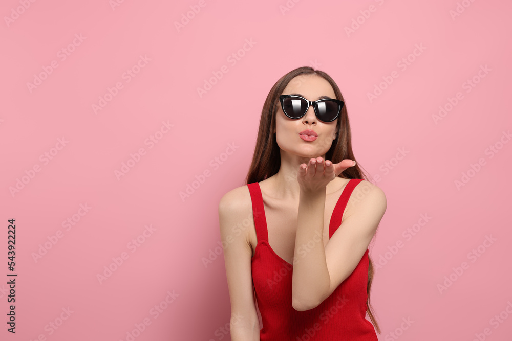 Beautiful young woman with sunglasses blowing kiss on pink background, space for text