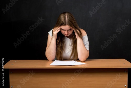 a woman of European appearance sits at a table with her hands behind her head over sheets of paper. tired teacher against the background of a slate wall. effects of stress