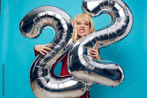 a woman in a red shirt stands showing her teeth on a blue background and holds inflatable balloons in the shape of the number twenty-two in silver color hugging them with her hands photo