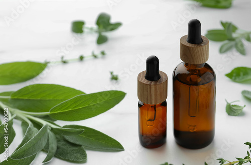 Bottles of essential oils and fresh herbs on white table  space for text