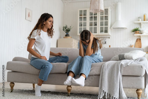 Strict woman looks at daughter sits on couch crying after ban on walks or home arrest. Little Caucasian girl was offended by mother after reprimand related to poor school performance or bad behavior. 