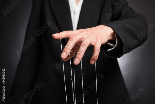 Woman in suit pulling strings of puppet on dark background, closeup photo