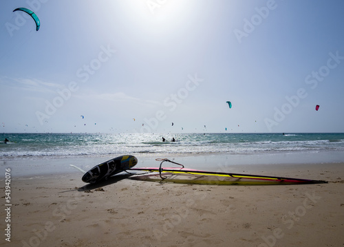 Beach on the coast of light with turquoise blue sea. Board with its sail waiting for windsurfing. In the background unrecognizable kite surfing.