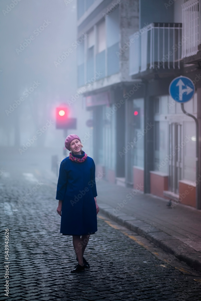 A blonde woman in an overcoat stands on the sidewalk in the morning fog.