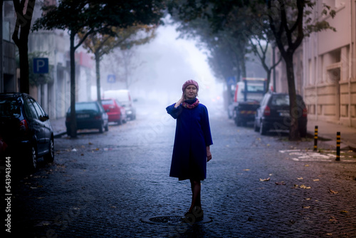 Portrait of a blond woman in an overcoat standing on a shady street.