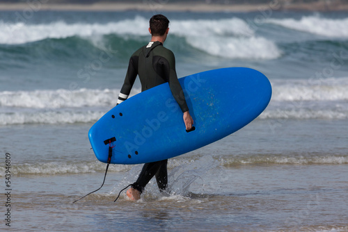 A surfer enters the water at A Lanzada beach with a blue apprentice surfboard