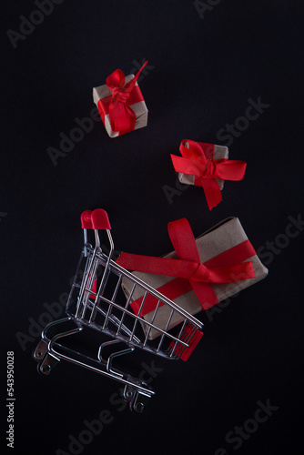 Happy shopping. Black friday holiday. Gift boxes with red ribbons fall in shopping cart. Black background, vertical photo