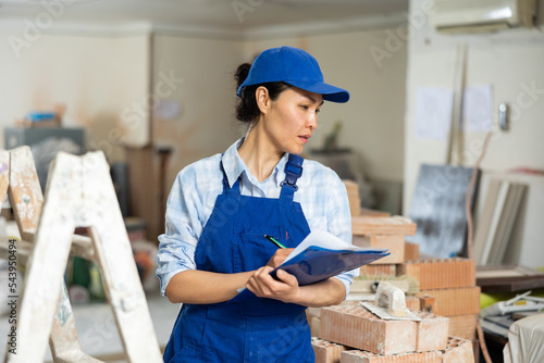 Portrait of confident woman builder planning work process for renovating object, making notes at clipboard