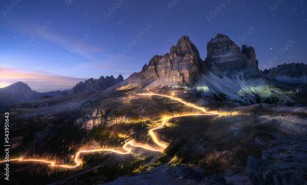 Car light trails on mountain road and high rocks at night in autumn in Tre cime, Dolomites, Italy. Colorful landscape with blurred light trails, hills, mountain peaks, sky with stars in fall at sunset