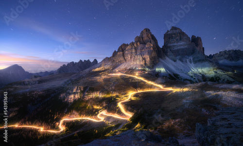 Car light trails on mountain road and high rocks at night in autumn in Tre cime, Dolomites, Italy. Colorful landscape with blurred light trails, hills, mountain peaks, sky with stars in fall at sunset