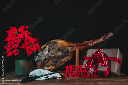 Spanish ham with a poinsettia, a gift, a merry christmas sign and the knife on a black background with a gift box.