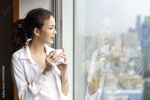 Portrait of confident and beauty young asian businesswoman with coffee cup standing and looking away in modern hotel with city blurred background. Success business and lifestyle concept