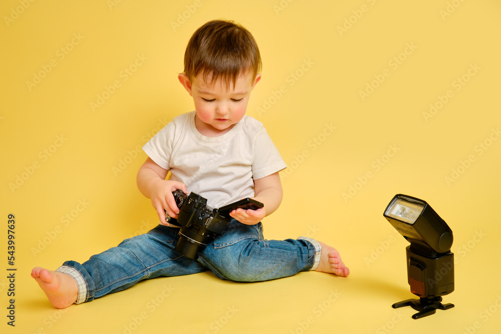 Toddler baby photographer with a camera on a studio yellow background. Happy child with photo equipment in a white t-shirt and blue jeans. Kid aged one year and four months Toddler baby photographer with a camera on a studio yellow background. Happy child with photo equipment in a white t-shirt and blue jeans. Kid aged one year and four months