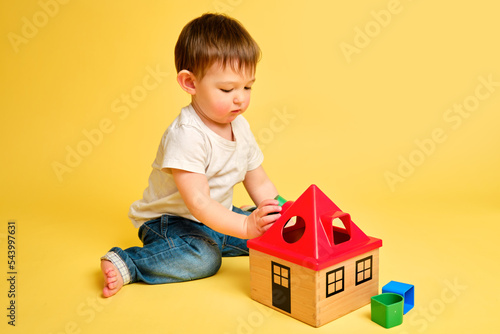 Toddler baby is playing sorter logic educational games with geometric shapes on a studio yellow background. Happy child play with educational toy house, learning logic. Kid aged one year four months photo