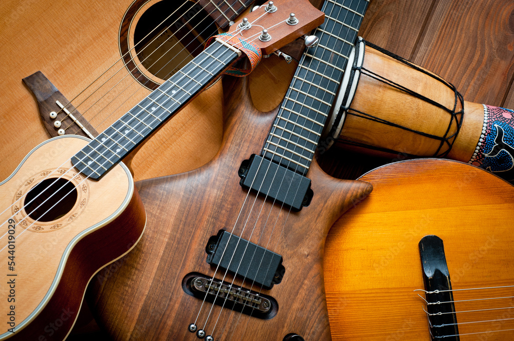Acoustic and electric guitars, ukulele and djembe standing side by side on a wooden background. Acoustic and electric guitars, ukulele and djembe standing side by side on a wooden background.