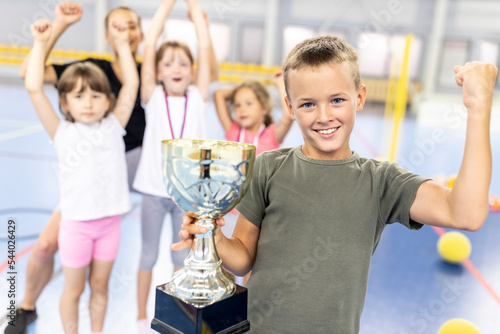 Happy boy flexing muscle holding trophy with friends at school sports court photo