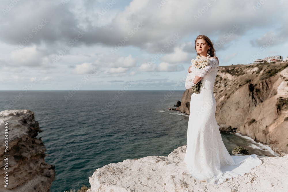 Romantic bride, blond girl in white wedding dress with open shoulders posing with sea and rocks in background. Stylish young woman standing on cliff's edge and looking into a ocean mountains at sunset Romantic bride, blond girl in white wedding dress with open shoulders posing with sea and rocks in background. Stylish young woman standing on cliff's edge and looking into a ocean mountains at sunset