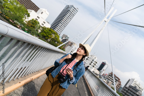 dutch angle shot of cheerful asian female visitor admiring the beauty of downtown Osaka city while walking on a bridge in Japan against blue sky