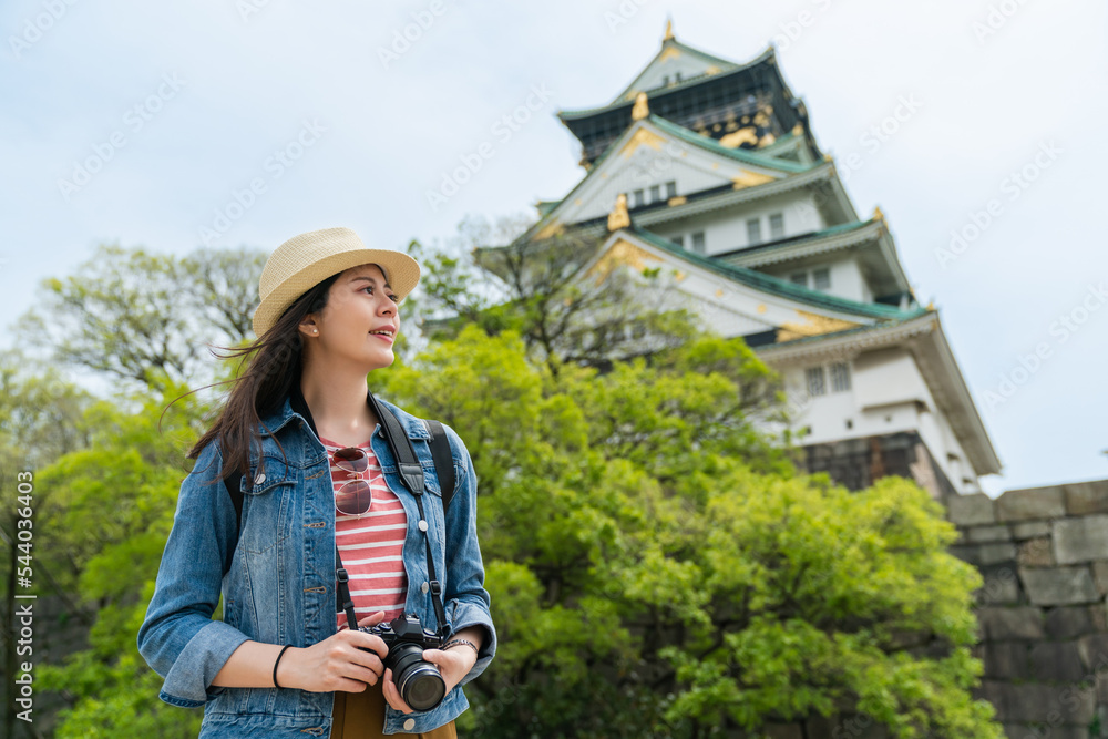 low angle shot portrait of happy asian girl photographer enjoying breeze and looking into distance with magnificent Osaka castle at background in japan