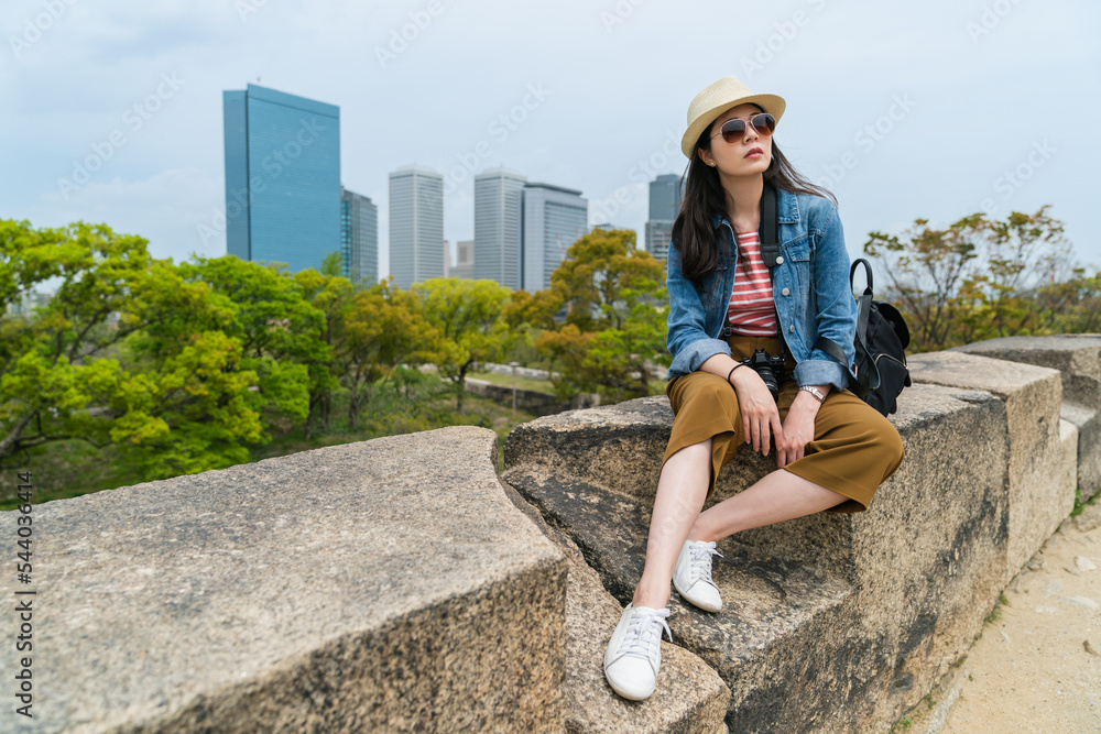 fashionable korean female traveler in hat and sunglasses sitting resting on rock and looking into distance with skyscrapers at background while visiting Osaka castle in japan