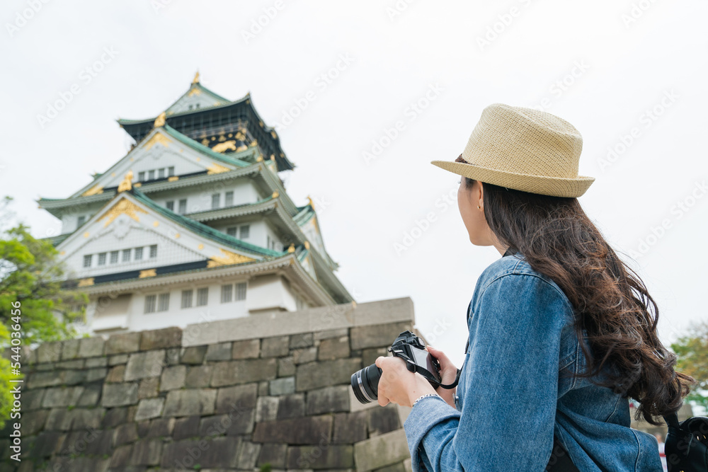 rear view of Asian woman photographer wearing hat looking and taking photo of beautiful Osaka castle with digital camera in japan.