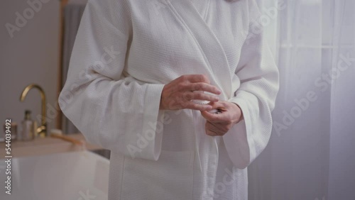 Close up cropped view unrecognizable old Caucasian woman unknown senior mature lady female in bathroom after bathing hygiene procedure in bath ties belt on white soft cozy comfortable bathrobe cloth