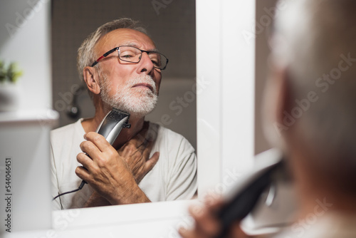 Foto Senior man is trimming his beard with electric trimmer in bathroom