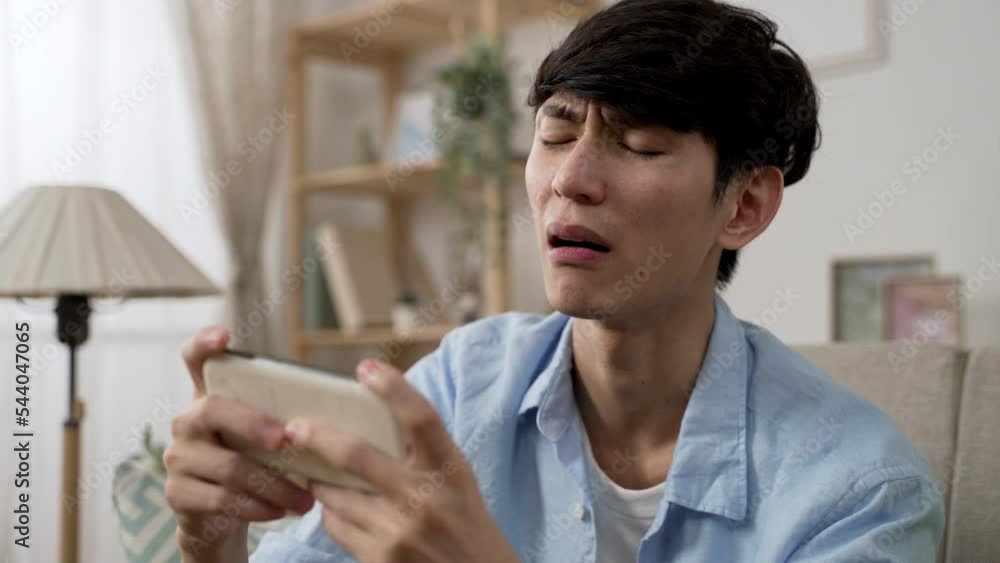 frustrated asian man feeling upset about losing the game he is playing on smartphone in the living room at home during daytime frustrated asian man feeling upset about losing the game he is playing on smartphone in the living room at home during daytime