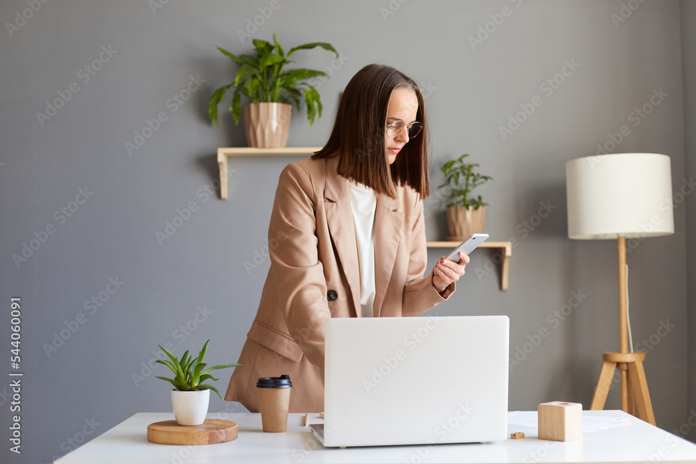 Portrait of dark haired woman freelancer standing near her workplace with notebook and holding smart phone in hands, calling to her partner, dialing number.