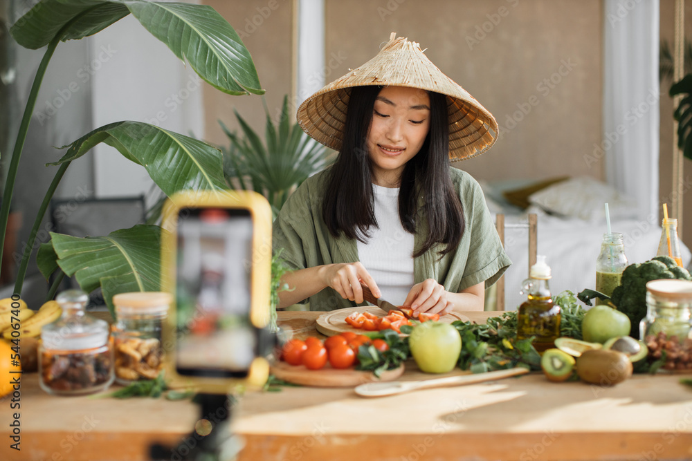 Young asian woman blogger or content creator chopping tomatoes preparing vegan salad and recording video on phone. Attractive female showing how cook healthy food while recording blog with smartphone