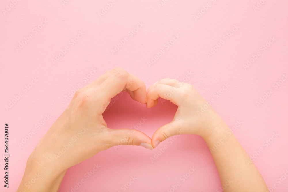Fototapeta premium Heart shape created from little daughter and young mother hands on light pink table background. Pastel color. Lovely emotional, sentimental moment. Love, happiness and safety concept. Top down view.