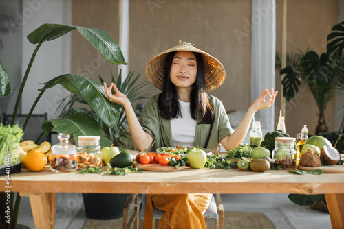 Fototapeta Young asian woman sitting near table with organic vegetables in yoga position while making salad, enjoying healthy diet, indoor at light exotic kitchen studio interior