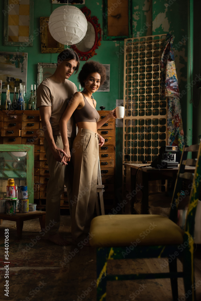 sexy african american woman with cup of tea holding hands with boyfriend and smiling at camera in art studio.