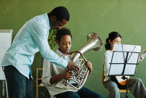 Young confident African American teacher of music consulting diligent schoolboy with trumpet against pre-teen girl playing guitar photo