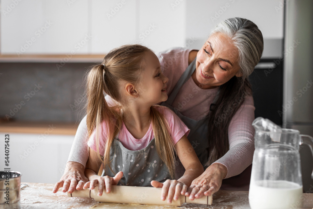Cheerful pretty caucasian little granddaughter and elderly grandmother tell dough with rolling pin for baking Cheerful pretty caucasian little granddaughter and elderly grandmother tell dough with rolling pin for baking