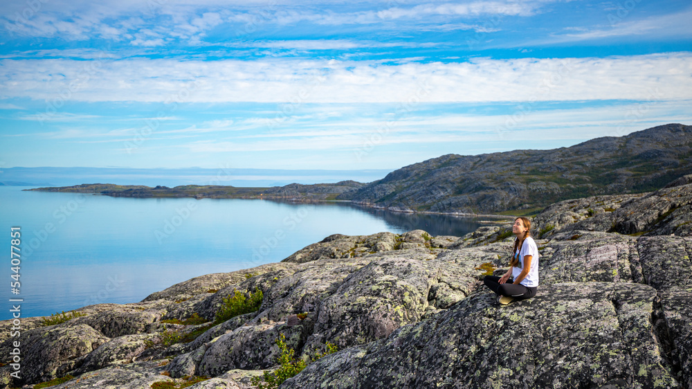 girl sits on rocks admiring the sunrise over a bay in northern norway; hiking in nordic norway, rugged landscape of the norwegian fjords