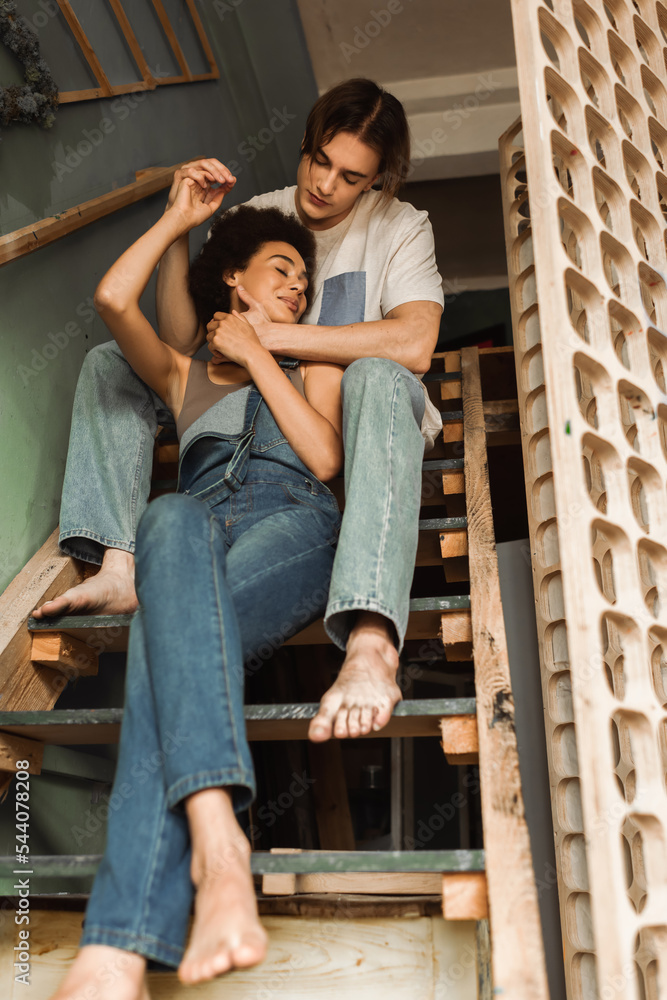 low angle view of barefoot african american woman in denim overalls holding hands with boyfriend on stairs in workshop. low angle view of barefoot african american woman in denim overalls holding hands with boyfriend on stairs in workshop.